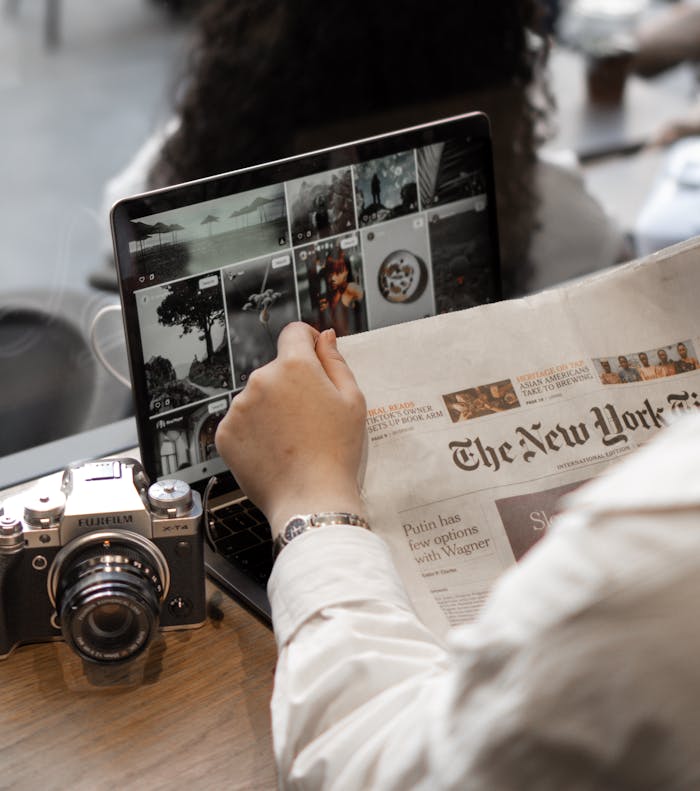 gallery-4 A person multitasks by reading a newspaper and using a laptop, surrounded by a vintage camera in a cafe setting.