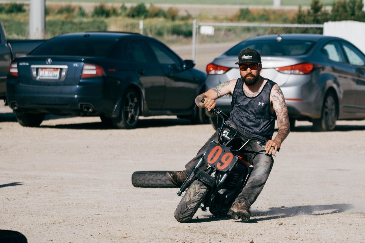about-us-01 A tattooed man riding a small motorcycle performs a stunt in a sunny parking lot.