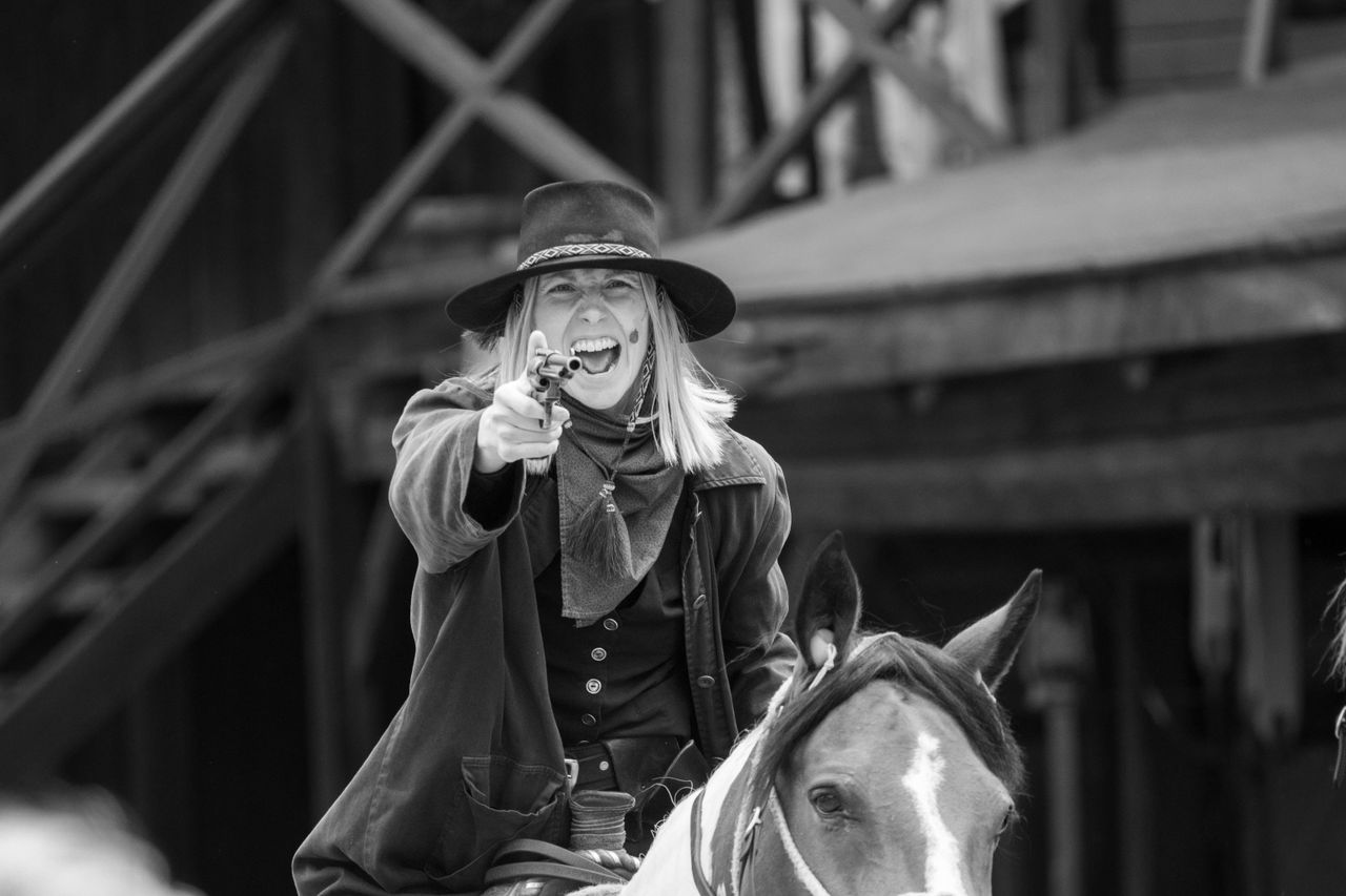 Grayscale photo of a woman in a black hat aiming a gun while riding a horse in a western setting.